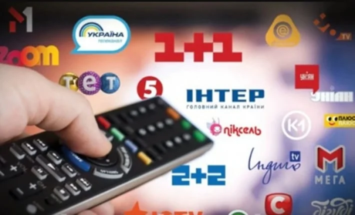 Рука тримає пульт навпроти екрана з логотипами українських телеканалів